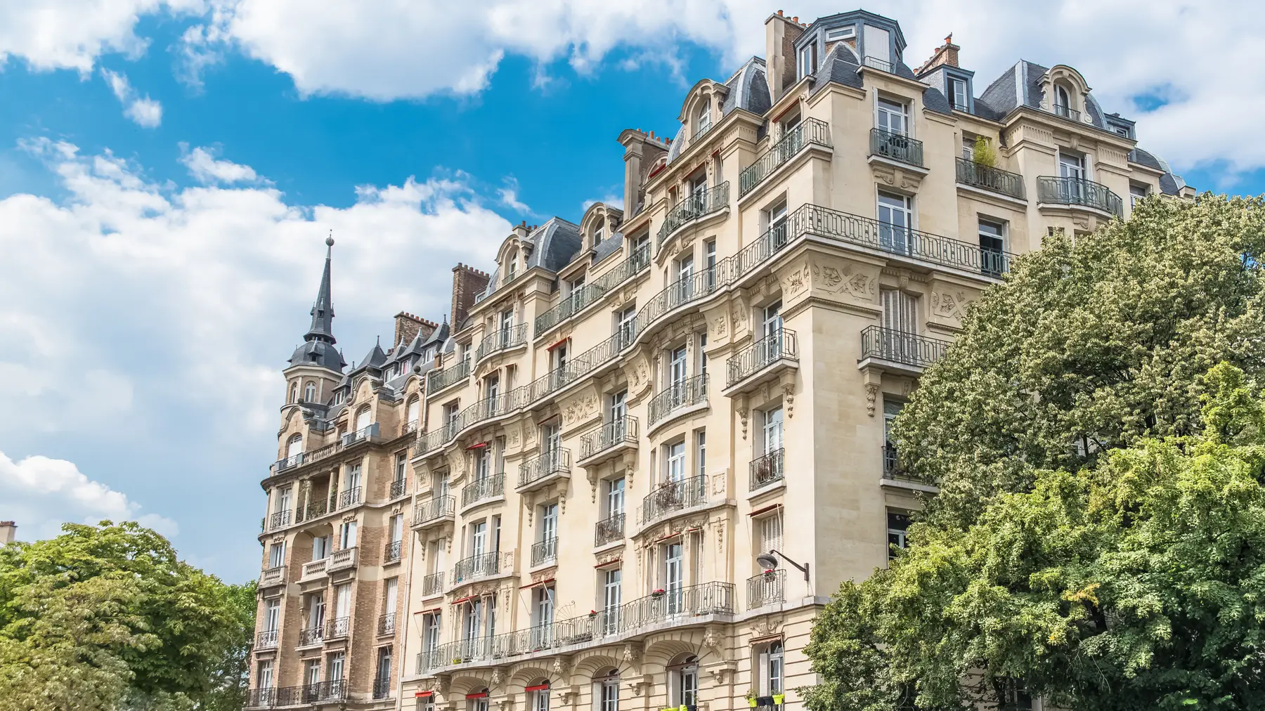 A Haussmann facade in full sun, stone balconies and mansard roof against a blue sky.